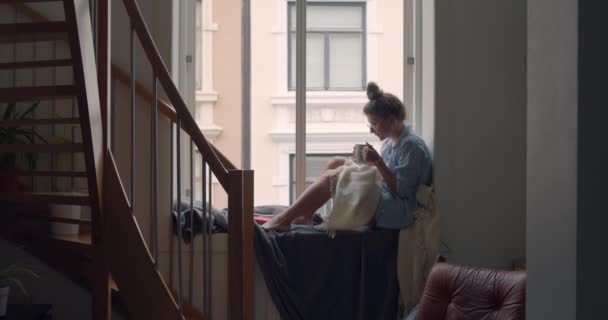 Молодая женщина в уютной пижаме пьет кофе на балконе — стоковое видео