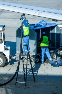 Havaalanının bir çalışanı tankeri çalıştırıyor ve uçağı havacılık yakıtıyla dolduruyor. Diğer işçiler bavulları uçağa yükler..