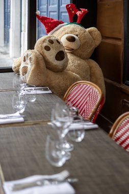 Noel temalı saç bandı takan büyük bir oyuncak ayı Paris 'teki bir kafenin terasında bir masada oturuyor. Masalar şarap kadehleriyle dolu, müşteriler için hazır..