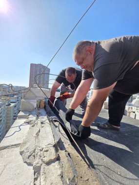 Kyiv, Ukrayna - Sep 5, 2025: Tanınmayan iki işçi bir binanın tepesindeki su geçirmez bir çatıyı onarıyor. Şehrin ufuk çizgisi arka planda mavi bir gökyüzünün altında görünür..