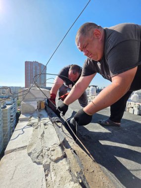 Kyiv, Ukrayna - Sep 5, 2025: Tanınmayan iki işçi bir binanın tepesindeki su geçirmez bir çatıyı onarıyor. Şehrin ufuk çizgisi arka planda mavi bir gökyüzünün altında görünür..