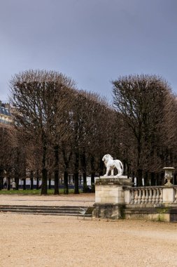Fransa 'nın Paris kentindeki Lüksemburg Bahçeleri' ndeki çakıl yolunda bir taş üste beyaz bir aslan heykeli bulunuyor. Bulutlu bir sonbahar gününde arka planda çıplak ağaçlar ve insanlar var..