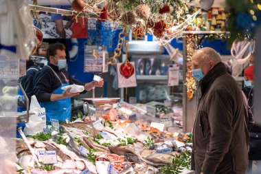 Paris, Fransa - 4 Aralık 2021: Mavi önlüklü bir satıcı ve deniz ürünleri tezgahında maske takan bir müşteri. Balıklar ve kabuklu deniz ürünleri fiyat etiketleriyle sergileniyor ve arkada Noel süslemeleri bulunuyor.