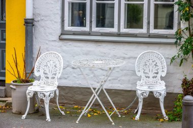 Almanya 'nın kuzeyinde, ortaçağ Lubeck, Holstein eyâletinde, binalar ve sonbahar yapraklarıyla birlikte tarihi bir kaldırımda metal bir zincirle zincirlenmiş eski tip demir masa ve sandalyeler..