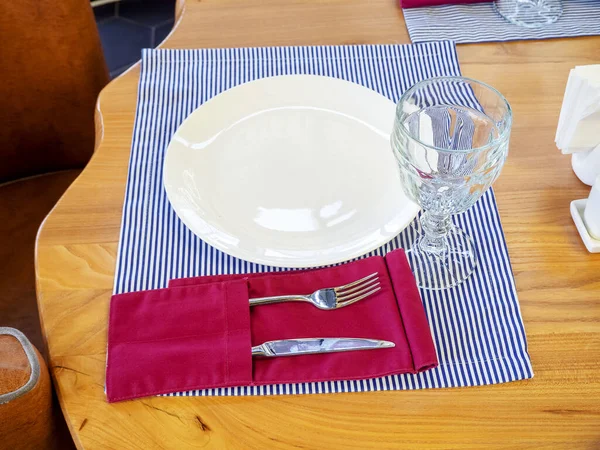 Een wit porseleinen bord met transparant glas en zilverwerk liggen op een gestreepte servet op een houten tafel. Tafelopstelling in een café. Close-up foto — Stockfoto