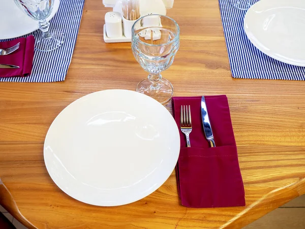 Een witte plaat met een kristalglas en zilverkleurig bestek in een rood servet op tafel. Cafe tafel gedekt. Close-up foto — Stockfoto