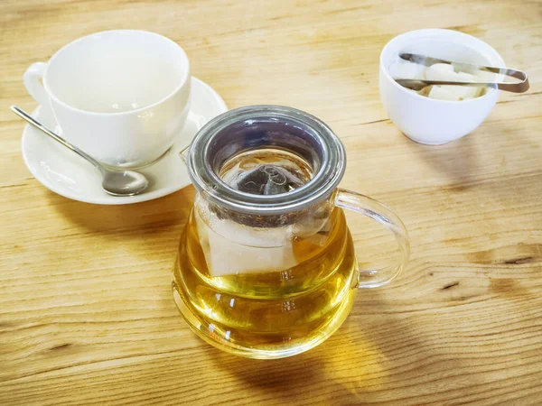一个透明的玻璃杯茶壶，茶壶旁边是一个装有糖和干净杯子的花瓶，放在木制表面上。茶党 — 图库照片