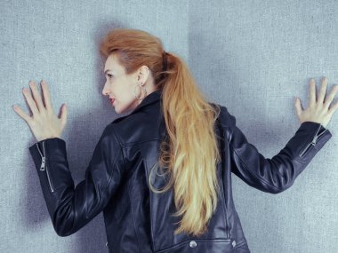 Siyah deri ceketli bir kız, gri duvar kağıdıyla duvara yaslanmış.