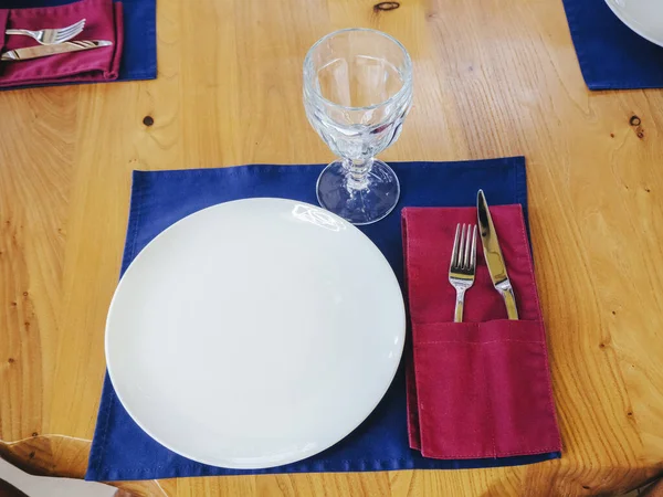 Witte plaat met een glazen vork en mes op een blauw servet op een houten lichttafel. Nieuw glanzend bestek. Close-up foto — Stockfoto