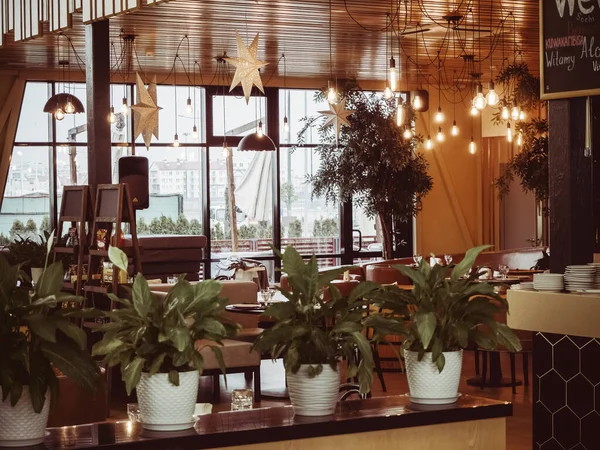 Russia, Sochi 14.03.2020. L'interno del caffè con le piante in piedi su uno stand in primo piano, molte lampade decorative luminose su un soffitto di legno e un'ampia finestra sul pavimento sullo sfondo — Foto Stock