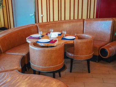 Yumuşak yarı dairesel deri koltuk, iki yuvarlak deri sandalye ve üzerinde ahşap bir masa ve çatal bıçak takımı olan bir masa.