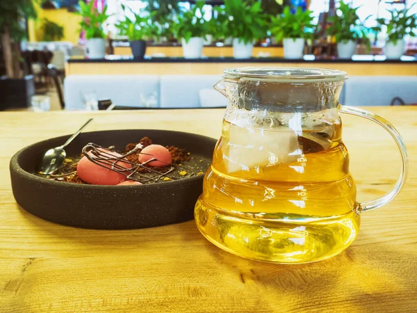 Прозрачный кувшин с зеленым чаем и розовый десерт в голубой тарелке на деревянном столике в кафе — стоковое фото