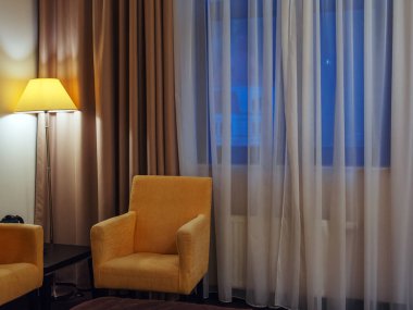 Pencerenin yanındaki odanın köşesinde, parlak bir lambanın yanında sarı bir koltuk. Bir otel odasının iç kısmının parçası.