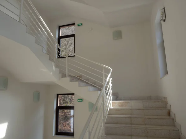 A stairwell with light steps with white railings and white walls and windows in dark frames