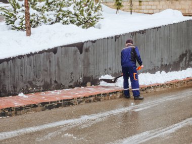 Mavi üniformalı bir hademe karları kaldırımdan kürekle kaldırıyor.