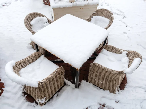 A square table with four wicker rattan chairs strewn with snow stands on a snow-covered tile — Stock Photo, Image