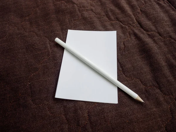 Uma folha de papel branca com um lápis branco jaz sobre um fundo marrom drapeado — Fotografia de Stock