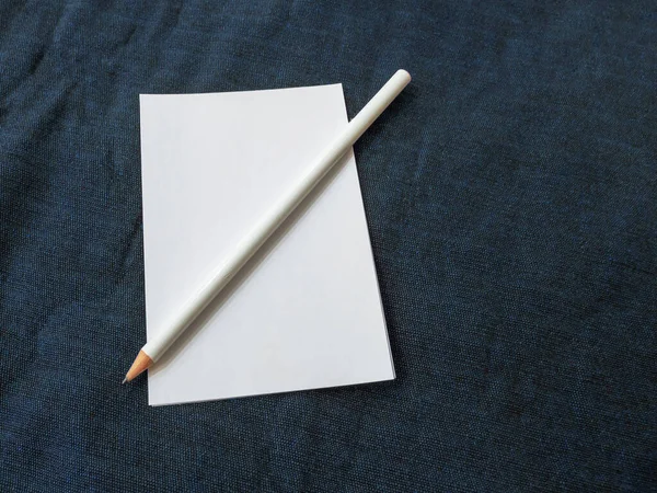 Uma folha de papel branca com um lápis de grafite branco fica em uma superfície de tecido azul — Fotografia de Stock