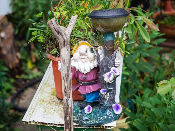 A ceramic statuette of a gnome stands next to a wooden pole on a background of green plants — Stock Photo, Image