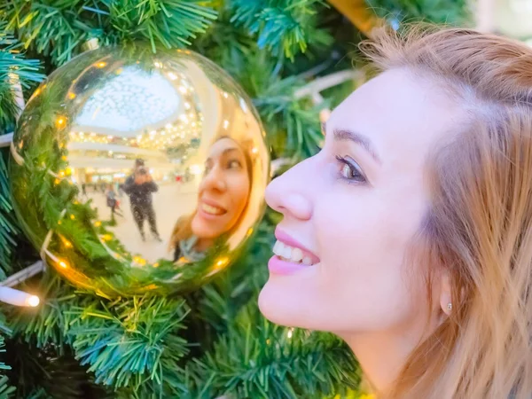 Smiling female profile near a golden ball with a reflecting face in it against the background of a christmas tree