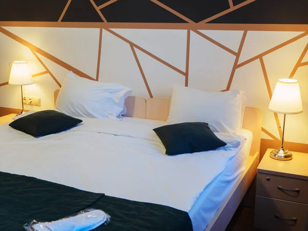 A neat double bed with lit floor lamps on the nightstands and a geometric pattern on the wall. Hotel room