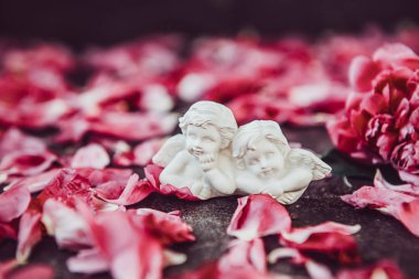 Statuette of two antique little lovely angels of the gypsum on the black stone background with pink peony flower petals. Vintage Love, romantic, Valentines day concept. Close up, soft selective focus clipart