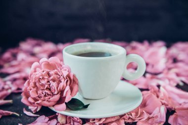 Bardak taze demlenmiş kahve ve pembe peony çiçek yaprakları ile siyah arka plan üzerinde uçan daire kapatın. Günaydın kavramı. Sessiz renkler. Vintage tonlama. Yumuşak seçici odak, kopya alanı