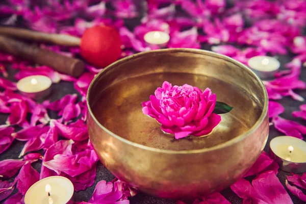Tibetan singing bowl with floating inside in water purple peony flower. Burning candles, special sticks and petals on the black stone background. Meditation and Relax. Exotic massage. Selective focus