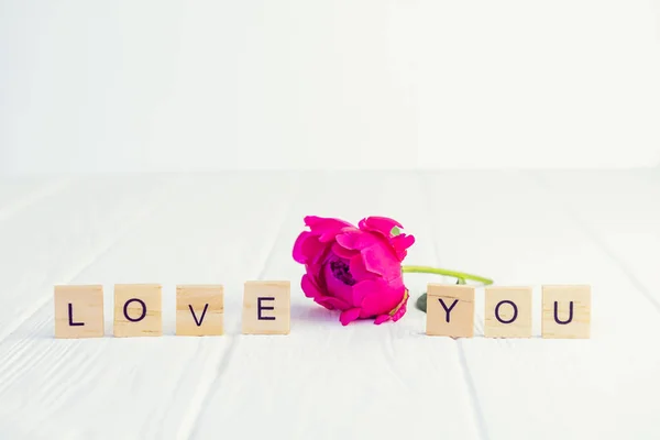 Message I love you spelled in wooden blocks with pink peony bud flower on white wooden table. Love, Valentines day background. Gift, greeting, compliment concept. Selective focus. Copy space