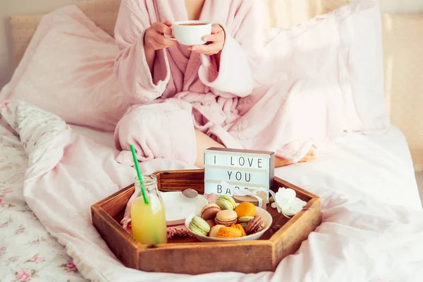 Romantic Breakfast in bed with I love you text on lighted box, macaroons, gift box on wooden tray and blurred cropped woman in a bathrobe drinking her coffee. Birthday, Valentine\'s day morning