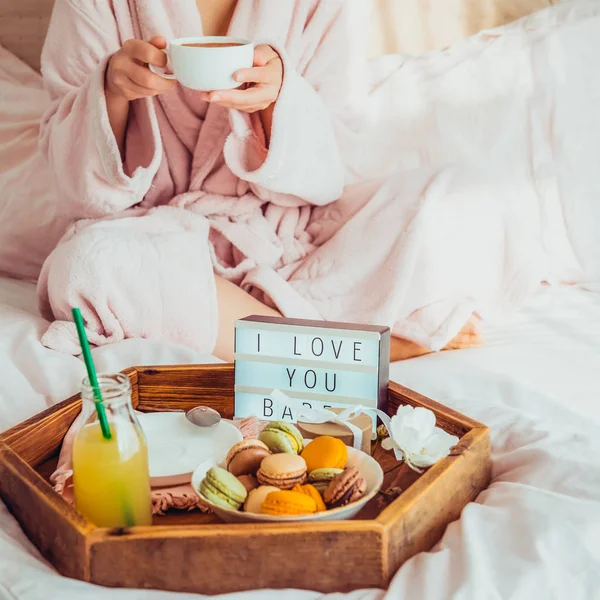 Romantic Breakfast in bed with I love you text on lighted box, macaroons, gift box on wooden tray and blurred cropped woman in a bathrobe drinking her coffee. Birthday, Valentine's day morning. Square.