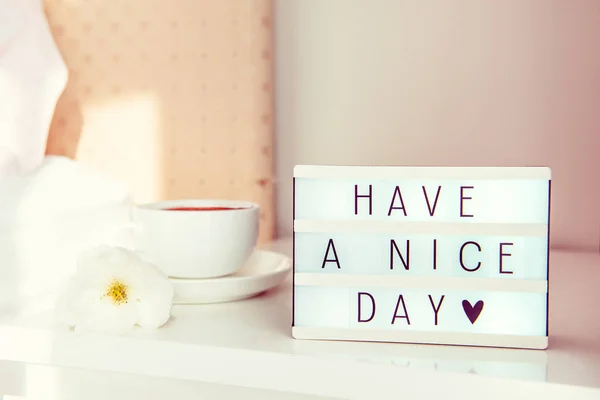 Have a nice day text message on lighted box, cup of coffee and white flower on the bedside table in sun light. Good morning mood. Hospitality, care, service concept. Selective focus. Copy space