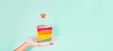 Birning mum ile Gökkuşağı pasta dilimi ile beyaz plaka üzerinde mavi arka plan izole yıldız şeklinde tutan el. Mutlu yıllar, parti kavramı. Geniş afiş. Seçici odak. Kopya alanı