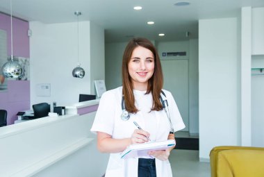 Kameraya bakarak ve neşeyle modern klinik receptional salonunda poz gülümsüyor Not defteri ile genç kadın doktor portresi. Sağlık ve Tıp. Kopya alanı