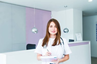 Kameraya bakarak ve neşeyle modern klinik receptional salonunda poz gülümsüyor Not defteri ile genç kadın doktor portresi. Sağlık ve Tıp. Kopya alanı