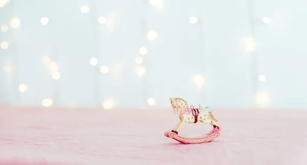 Figura de porcelana de juguete árbol de Navidad vintage de un caballo mecedora de pie sobre el mantel rosa sobre el fondo de luces borrosas. Disposición festiva. Amplio estandarte. Enfoque selectivo suave. Copiar espacio — Foto de Stock