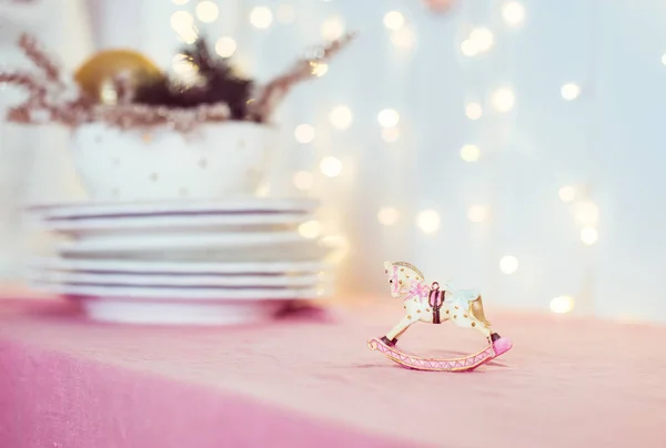 Composición con árbol de Navidad vintage juguete porcelana figurita de un caballo mecedora y platos en la mesa rosa en el fondo de luces borrosas. Disposición festiva. Enfoque selectivo suave. Copiar espacio . — Foto de Stock