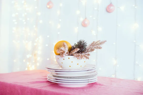 Christmas table setting. Not served white dishes with gold decor and fir-tree on pink tablecloth with blurred lights and new year balls on wall. Festive background. Soft selective focus. Copy space. — Stock Photo, Image