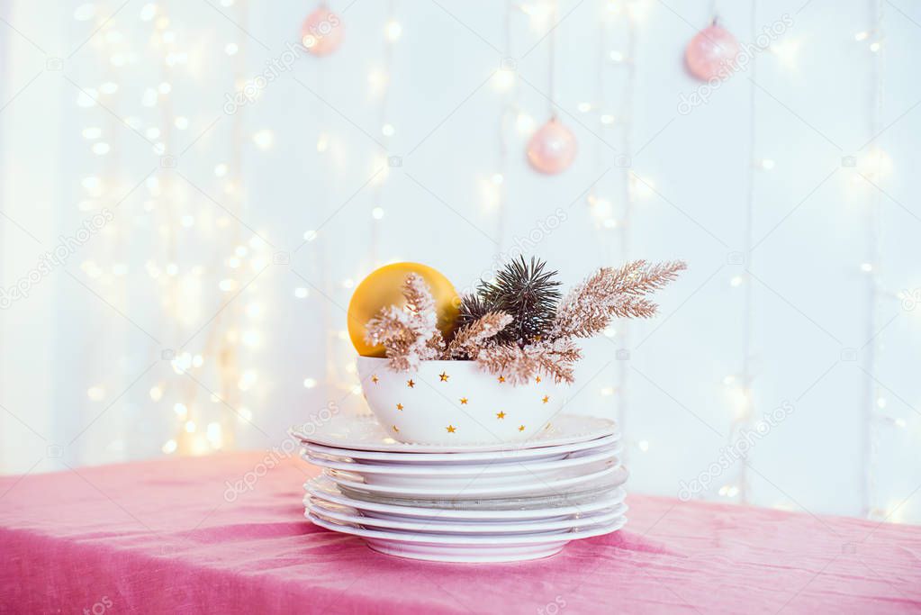 Christmas table setting. Not served white dishes with gold decor and fir-tree on pink tablecloth with blurred lights and new year balls on wall. Festive background. Soft selective focus. Copy space.