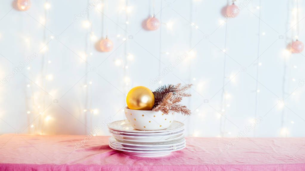 Christmas table setting. Not served white dishes with gold decor and fir-tree on pink tablecloth with blurred lights on the wall. Festive background. Wide banner. Soft selective focus. Copy space.