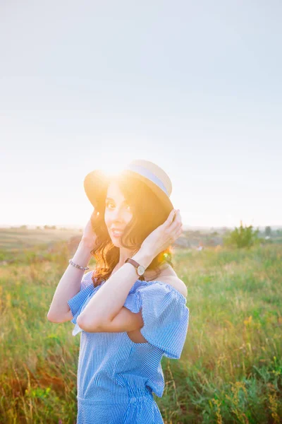 Portret van jonge vrouw in blauwe romantische jurk en stro hoed op groene weide genieten van de zomer zonsondergang. Rust en harmonie. Zomer vakantie, leuk, positieve stemming. Terug zon licht, zonnestralen. Ruimte kopiëren. — Stockfoto