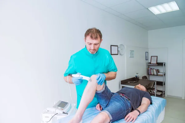 Homem jovem que recebe o laser ou a massagem de terapia de ímã em um joelho a menos dor. Um quiroprático trata a articulação do joelho do paciente em consultório médico. Neurologia, osteopatia, quiropraxia. Espaço de cópia . — Fotografia de Stock