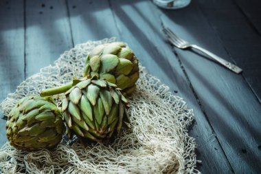 Kapatın Siyah ahşap masa üzerinde örgülü ip ve mutfak şeyler ile taze yeşil enginar kapatın sabah pencere ışığı ile aydınlatılmış. Çiftçi hasadı, yerel pazar. Seçici odaklama, Boşluk kopyalama.