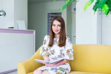 Tam büyüme Portre Gülümseyen genç iş kadını iş salonunda sarı kanepede oturan ve not defterine yazma, yararlı bilgiler notlar yapar, zarif giyinmiş. Kopyalama alanı.