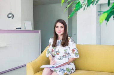 Tam büyüme Portre Gülümseyen genç iş kadını iş salonunda sarı kanepede oturan ve not defterine yazma, yararlı bilgiler notlar yapar, zarif giyinmiş. Kopyalama alanı.