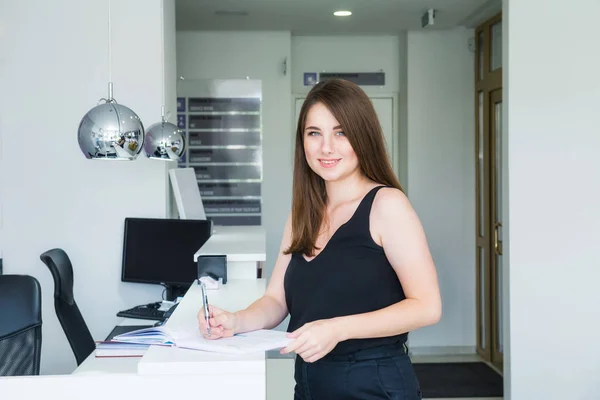 Retrato de Sorrindo jovem gerente do sexo feminino em pé na recepção na sala de escritório e escrevendo em caderno, faz anotações de informações úteis, vestidas casualmente. Foco seletivo, espaço de cópia . — Fotografia de Stock