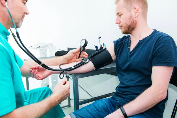 Médico masculino usando esfigmomanómetro con estetoscopio comprobando la presión arterial a un paciente varón joven en el hospital. Atención médica, estilo de vida saludable y concepto de servicio médico. Copiar espacio . —  Fotos de Stock