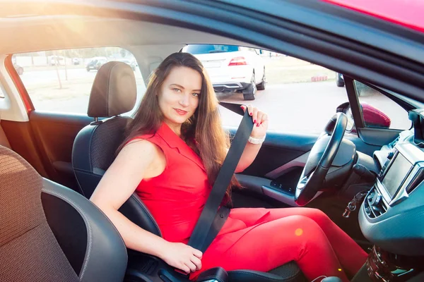笑顔のビジネス女性の肖像画、赤い夏のスーツを着た白人の若い女性ドライバーは、カメラを見て、ホイールカーの後ろに座っている間、彼女のシートベルトを着用します。選択的な焦点、コピースペース. — ストック写真