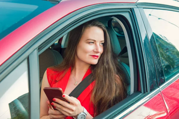 Молодая белая женщина-водитель в красном костюме держит свой смартфон и смотрит в сторону, сидя за рулем красной машины. Выборочный фокус, копировальное пространство . — стоковое фото