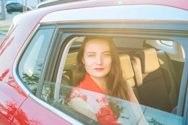 Porträt einer jungen kaukasischen Frau im roten Anzug, die durch das Fenster in die Kamera blickt und im Abendsonnenlicht auf dem Rücksitz im Auto sitzt. Selektiver Fokus, Kopierraum. — Stockfoto